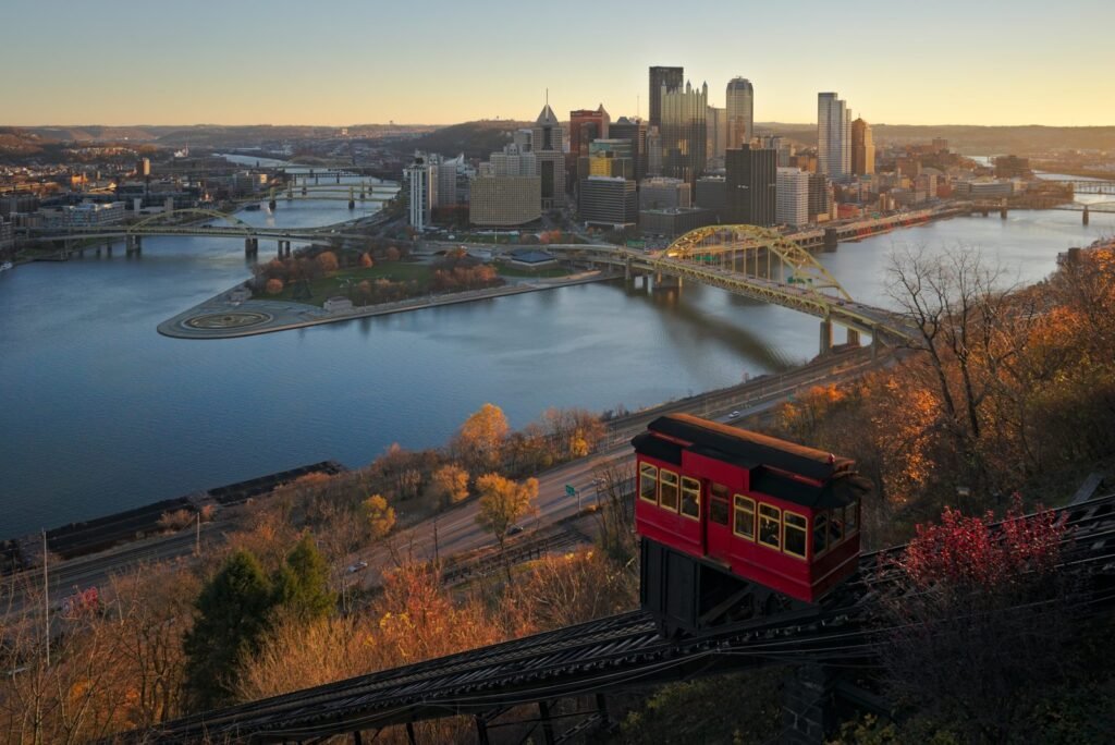 A red Duquesne Incline car climbing Mount Washington above the Pittsburgh skyline and the confluence of the Allegheny and Monongahela rivers in autumn